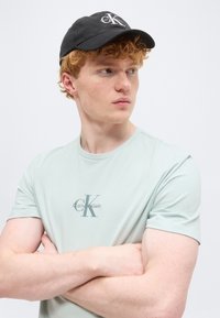 Άνδρας με κόκκινα μαλλιά, με σταυρωμένα χέρια, φορώντας ένα ανοιχτό πράσινο T-shirt Calvin Klein και ένα μαύρο καπέλο Calvin Klein, κοιτάζοντας προς τα αριστερά του.