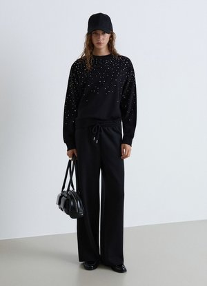 Donna con berretto nero, maglione nero con piccoli puntini bianchi, pantaloni neri larghi, tiene una borsa nera, in piedi davanti a uno sfondo bianco uniforme.