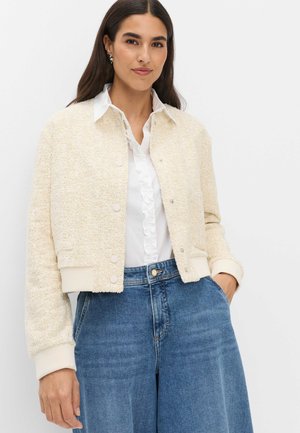 Vrouw draagt een crèmekleurig gestructureerd jasje met knopen over een witte blouse met ruches en blauwe hoog opgebonden spijkerbroek, staand met één hand in de zak.