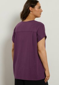 Lila Kurzarmoberteil mit lässiger Passform, ausgestattet mit einer Naht über den Rücken und aus glattem, leichtem Stoff. Flacher Saum, schlichtes Design.