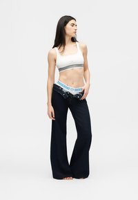 Γυναίκα με μακριά σκούρα μαλλιά που φοράει λευκό crop top και σκούρο τζιν με ευρείς παντελόνια, μερικώς ξεκουμπωμένο για να αποκαλύψει εσώρουχο με μοτίβο στη ζώνη.