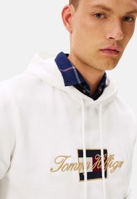 Sudadera blanca con cordones ajustables, que presenta un logo bordado en dorado de "Tommy Hilfiger" en un parche rectangular azul marino, rojo y blanco.