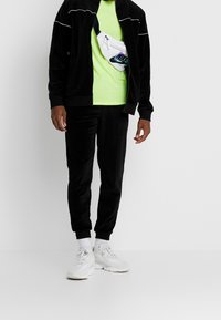 Schwarzer Velours-Trainingsanzug mit weißen Paspeln, kombiniert mit einem neon grünen T-Shirt und einer weißen Bauchtasche. Weiße Sneaker mit strukturierten Sohlen.