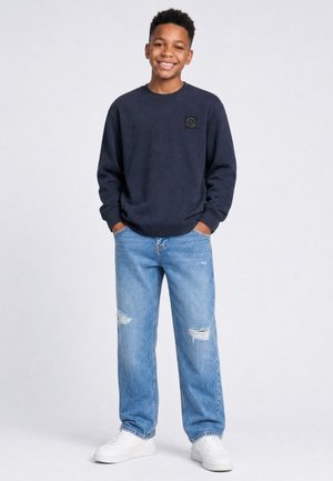 Lachende jongen staand met handen in de zakken, gekleed in een donker sweatshirt, gescheurde blauwe spijkerbroek en witte sneakers tegen een lichte achtergrond.