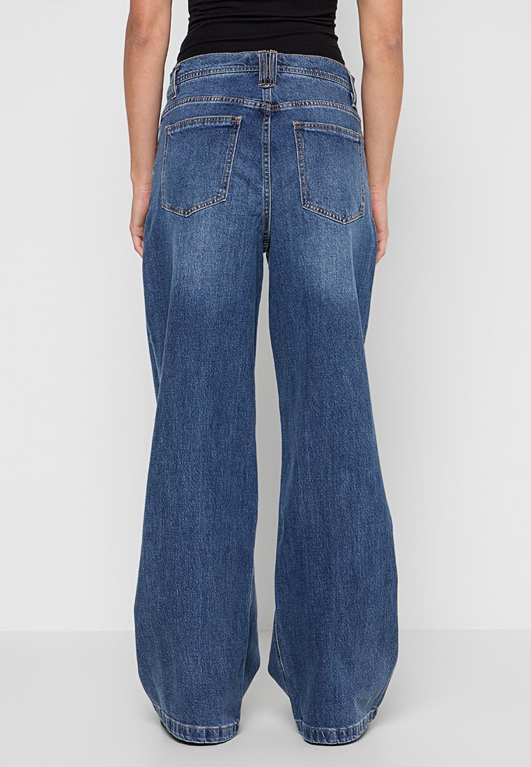 Person, die hoch sitzende, weit geschnittene blaue Jeans und ein schwarzes ärmelloses Oberteil trägt, von hinten vor einem schlichten Hintergrund gezeigt.