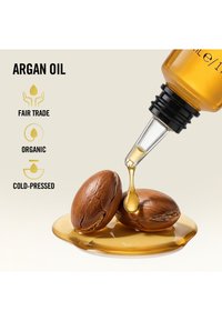 Cuentagotas soltando aceite de argán dorado sobre dos nueces de argán agrietadas con etiquetas de comercio justo, orgánico y prensado en frío.