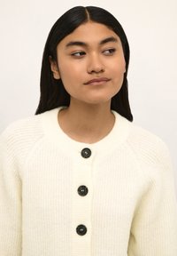 Krämfärgad, ribbad cardigan med rund halsringning och stora svarta knappar. Mjuk textur med en avslappnad passform, lämplig för lager-på-lager.