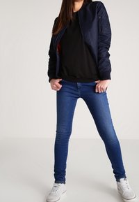 Námořnická bombardérka s žebrovanými manžetami, černé tričko a modré džíny slim-fit. Model s svetle šedými teniskami. Jednoduchý, neformální outfit.