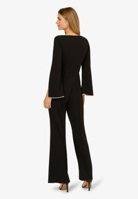 Černý overal s rozšířenými rukávy, zdobený perlovými akcenty na okrajích rukávů a přiléhavým siluetou, zobrazený ze zadní strany.