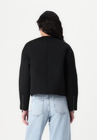 Jachetă neagră scurtă din țesătură texturată, cu guler înalt și manșete cu fermoar. Asortată cu blugi din denim de culoare albastră deschisă.