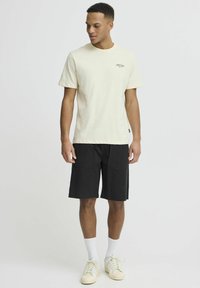 Svijetlosmeđi majica s kratkim rukavima i malim logom, uparen s crnim šortsevima koji imaju dvije zakrčaste džepove. Off-white tenisice upotpunjuju odjevnu kombinaciju.