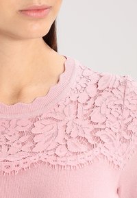 Pull en rose avec un empiècement en dentelle texturée, un col en forme de coquille et des poignets côtelés, orné de motifs floraux pour un détail supplémentaire.