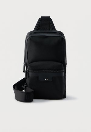 Sac bandoulière compact noir avec poche zippée à l'avant, sangle réglable et tissu texturé sur fond uni.
