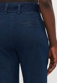 Mørkeblå denim bukser med en tett design, beltehemper og to baklommer med enkel søm. Glatt tekstur med en svak glans.
