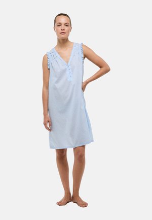 Femme debout pieds nus portant une robe bleu clair sans manches, longueur genou, avec des détails de boutons et des épaules volantées sur fond blanc.