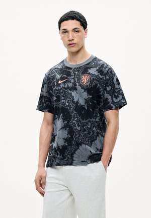 Jeune homme aux cheveux courts et foncés, portant un maillot de sport noir et gris à motifs avec des logos orange et un pantalon clair, debout avec une main dans la poche.