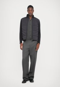 Man wearing a black quilted vest over a dark gray buttoned shirt with gray wide-leg trousers and black shoes, standing against white background.