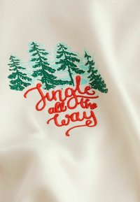 Tissu blanc avec une broderie d'arbres verts et la phrase « Jungle jusqu'au bout » en cursive rouge. Texture lisse avec des accents colorés.