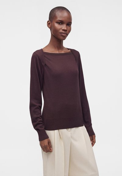 Femme aux cheveux courts portant un haut à manches longues violet foncé et un pantalon beige ample sur un fond uni.