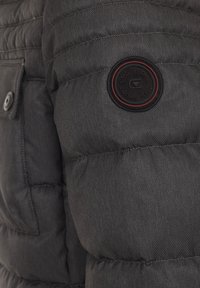Grå puffjacka med texturerad tyg, med en rund svart logotyp patch med röda accenter på vänster ärm och en knappad ficka.