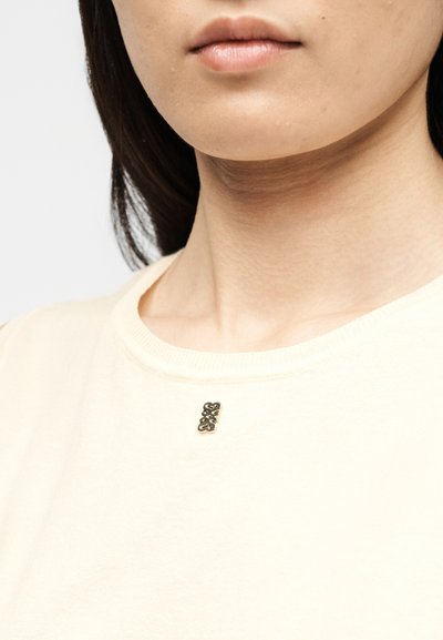 Κοντινό πλάνο στο κατώτερο μέρος του προσώπου και το λαιμό ενός ατόμου που φοράει κρεμ πουκάμισο με μια μικρή, περίπλοκη μεταλλική καρφίτσα προσαρτημένη κοντά στον γιακά.