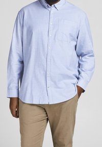 Camicia azzurro chiaro a maniche lunghe con colletto, taschino sul petto e bottoni bianchi, realizzata in un tessuto morbido e strutturato. Indossata con pantaloni beige.