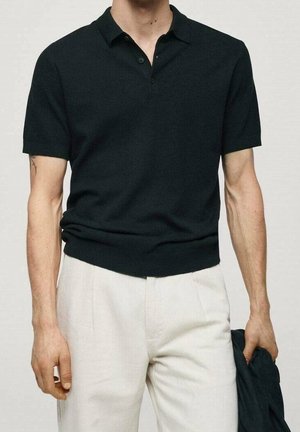 Mand iført en sort polo t-shirt med korte ærmer og hvide bukser, der holder en sort jakke i sin højre hånd.