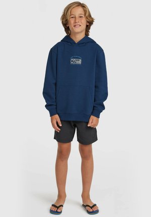 Tumši zils kapuces džemperis ar priekšējo kabatu un O'Neill logotipu. Valdāts kopā ar melniem šortiem un čībām. Audums šķiet mīksts un ikdienišķā stilā.