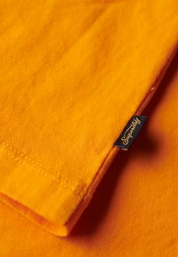 Tela de algodón naranja con una superficie texturizada, con un borde doblado y una pequeña etiqueta azul marino que dice "Superdry."
