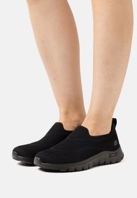 Black slip-on shoes made of textured knit fabric with a low-cut design and cushioned sole, featuring a branded logo on the side.