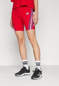 Personne portant un short rouge Adidas avec des rayures bleues et blanches, des chaussettes blanches Adidas avec des rayures noires, et des baskets noires Adidas sur un sol gris.