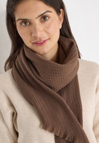 Écharpe marron texturée en matériau doux, avec un motif tissé et des extrémités frangées, portée sur un pull beige clair.