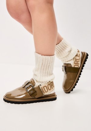 Smeđe lakirane cipele s kopčama, uparene s kremastim rebrastim čarapama s logotipom. Crna gumena potplat s teksturiranim gripom.