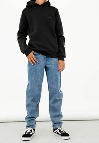 Sudadera negra con un bolsillo frontal, que presenta un pequeño logo cuadrado. Combinada con unos vaqueros de mezclilla azul sueltos y zapatillas negras con detalles blancos.