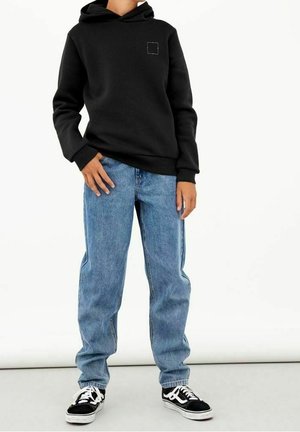 Sudadera negra con un bolsillo frontal, que presenta un pequeño logo cuadrado. Combinada con unos vaqueros de mezclilla azul sueltos y zapatillas negras con detalles blancos.