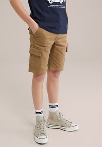 Kaki cargoshorts met twee zijzakken, gemaakt van katoen. Gekocht met grijze high-top sneakers en witte sokken met zwarte strepen.