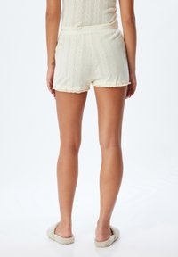 Pantalones cortos de color crema con textura, patrón acanalado y dobladillo deshilachado. Llevados con zapatillas de felpa, mostrando un estilo casual y cómodo.