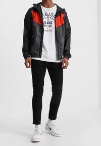 Veste coupe-vent noire avec des accents rouges sur les épaules, t-shirt graphique blanc, jeans noirs et baskets blanches, se tenant devant un fond uni.