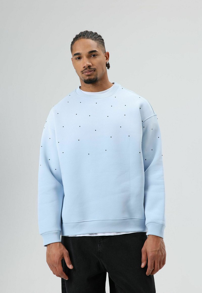 Sweatshirt bleu clair à col rond, avec des points noirs éparpillés, des poignets côtelés et une coupe légèrement oversize. Porté avec un jean foncé.