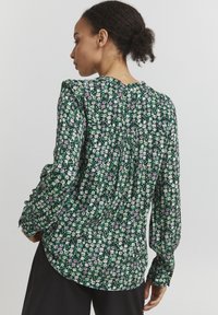 Blusa floreale verde con maniche lunghe, dettaglio arricciato sul retro e piccoli motivi di fiori bianchi e viola su un tessuto testurizzato.