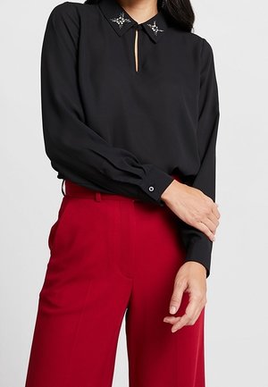 Femme portant un chemisier noir à manches longues avec un col orné et un pantalon rouge taille haute, debout les bras croisés à la taille.