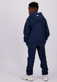 Giacca di jeans blu con cappuccio, vita elastica e pantaloni in jeans abbinati. Indossata con sneaker bianche. La texture sembra morbida.