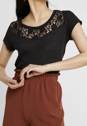Femme portant un haut en dentelle noire et un pantalon taille haute couleur rouille ajustant la ceinture avec les deux mains.