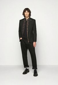 Svart dressjakke over en brun rutete skjorte, kombinert med svarte bukser og svarte sko. Stoffet virker glatt med strukturert skreddersøm.