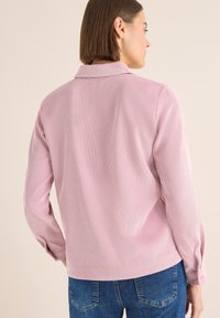 Camisa de pana a rayas rosas con cuello, mangas largas y puños abotonados; mostrada desde atrás sobre un fondo neutro.