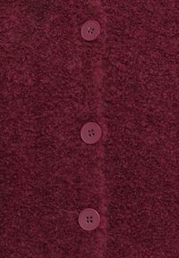 Detalhe de um tecido de lã texturizado em vermelho profundo com três botões redondos de quatro furos a combinar dispostos em coluna vertical.
