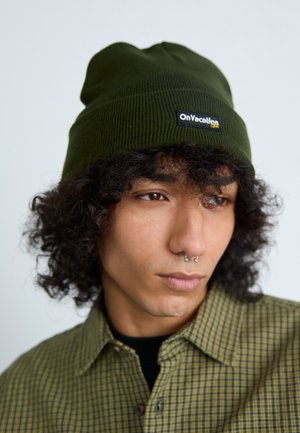 Roheline ribatud beanie musta sildiga, kus on kirjas "OnVacation" ja kollased aktsendid. Veidi lõtv sobivus, pehme tekstuur, kantud ruudulise särgiga.