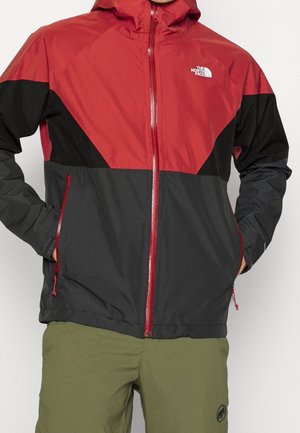 Mężczyzna w czerwonej, czarnej i ciemnoszarej kurtce z kapturem i zamkiem błyskawicznym, z logo The North Face na piersi, ręce w kieszeniach, ubrany w oliwkowe spodnie.