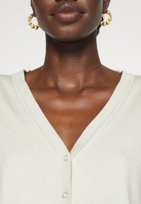 Gros plan sur le cou et le haut de la poitrine d'une personne portant des boucles d’oreilles créoles dorées et un cardigan crème à col en V avec des boutons en perle.