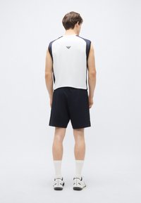 Biely tielkový top s námorníckymi modrými detailmi a logom, v kombinácii s čiernymi šortkami, nosený s bielymi ponožkami a športovou obuvou. Minimalistický dizajn.